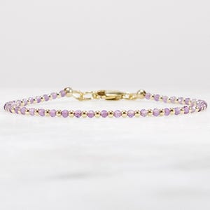 May include: Delicate bracelet featuring small, round, light purple beads interspersed with gold-colored beads. The bracelet has a gold-colored clasp and is displayed against a white background. A simple and elegant piece of jewelry.
