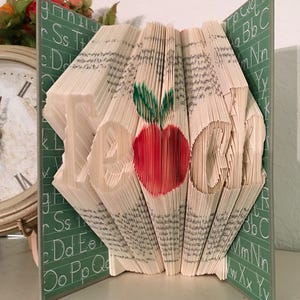 May include: A book folded into the shape of the word "TEACH" with a red apple and green leaf on top. The book is sitting on a green surface with white letters.