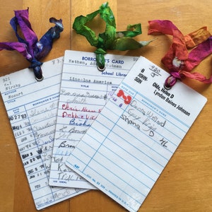 May include: Three vintage library cards with blue and white lines. The cards have handwritten text, including the titles "Lincolns America", "Mozart", and "Olds, Helen D Lyndon Baines Johnson". Each card has a hole punched in the top right corner and a colorful ribbon tied through the hole.