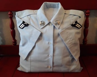 May include: A light blue pillow shaped like a military uniform shirt, complete with collar, buttons, and epaulettes. The pillow is resting on a red bench. The shirt has two US insignia on the collar.