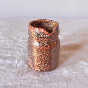 May include: A small, handcrafted ceramic creamer with a rustic aesthetic. The creamer features a brown and orange speckled glaze with a small spout and a reddish-orange rim. The cylindrical body has a slightly textured surface, suggesting a handmade quality.
