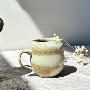 Può includere: Una tazza in ceramica fatta a mano con un corpo arrotondato e un manico curvo. La tazza presenta fasce orizzontali beige chiaro e marrone macchiettato. La tazza è su una superficie di colore chiaro, con un rametto di fiori bianchi sullo sfondo.