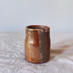 May include: A small, handmade ceramic pitcher with a rustic, textured finish. The pitcher features a warm color palette of orange, brown, and hints of green. It has a spout for easy pouring and is ideal for serving sauces or cream.