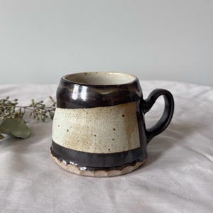 Pode incluir: Uma caneca de cerâmica com formato arredondado e alça, apresentando um design bicolor com uma faixa central em creme salpicado contrastada por seções esmaltadas em marrom escuro na parte superior e inferior, com uma aparência texturizada.