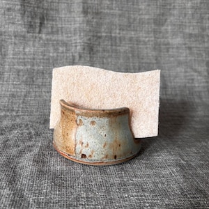 May include: A ceramic sponge holder with a mottled glaze in shades of tan, brown, and muted blue-green. The holder has a curved, low-profile design and contains a rectangular, light beige, fibrous cellulose sponge standing upright inside.