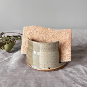 May include: A handmade ceramic sponge holder in a speckled, muted green glaze with a brown rim. A beige sponge is held in the holder. The holder is on a gray surface. Kitchen and home decor.