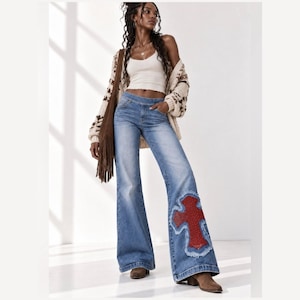Könnte beinhalten: Blaue Schlaghose aus Denim mit einer großen, roten Kreuz-Applikation am unteren linken Bein. Die Jeans hat eine mittlere Waschung, eine eng anliegende Taille und ein weites Bein. Das rote Kreuz ist mit ausgefransten Kanten und Nieten verziert.