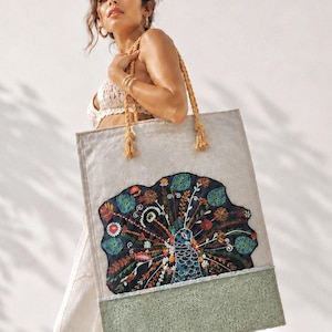 May include: A rectangular beige tote bag featuring a large, dark blue embroidered peacock motif with colorful floral and leaf details. The bag has thick, braided tan rope handles and a textured, light green panel along the bottom edge.