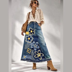 May include: A long denim skirt featuring a patchwork design with various blue fabric squares and large appliquéd flowers with yellow centers. The skirt has a frayed hemline and is paired with a long-sleeved cream-colored button-down shirt, a brown shoulder bag, and tan ankle boots.