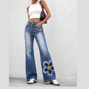 May include: A white sleeveless crop top and blue denim flare jeans. The jeans feature a high-waisted fit, a distressed rip on the right thigh, and a large dark blue flower appliqué with a leopard print centre on the lower left leg.