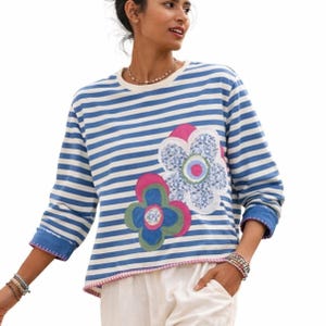 May include: A blue and white striped long-sleeve shirt with floral appliqués. The shirt features three-quarter sleeves with blue cuffs and a pink trim along the bottom. The floral designs include pink, blue, and green colors.