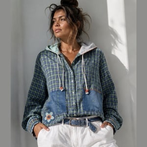 May include: A long-sleeve hooded shirt featuring a blue and green plaid pattern, a button-down front, and two large denim patch pockets. The shirt includes a light-colored hood lining, white drawstrings with orange accents, and a floral embroidery.
