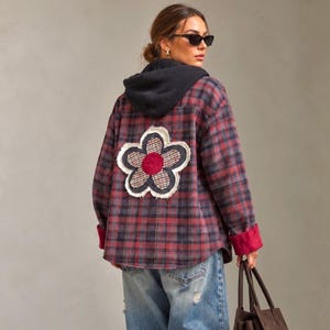 May include: A red and black plaid flannel shirt featuring a large, frayed-edge flower appliqué on the back with a red center and a checkered pattern. The shirt includes a dark gray ribbed hood and bright red cuffs on the long sleeves.