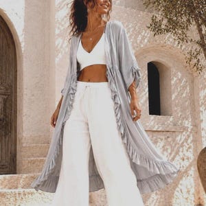 Pode incluir: Um kimono comprido azul claro, com frente aberta e bordas com babados. É combinado com um top curto branco e calças brancas de perna larga. O conjunto é leve e arejado, perfeito para climas quentes.