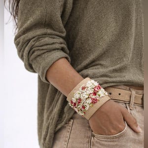 May include: A wide beige cuff bracelet decorated with small white and red fabric flowers. The bracelet is made of a woven material and is worn on a person's wrist. The person is wearing a green long-sleeved top and beige trousers.