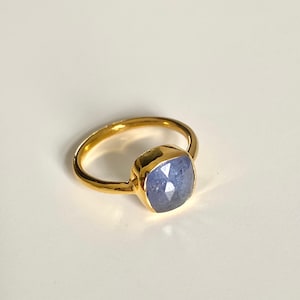 May include: A gold ring featuring a blue gemstone. The gemstone is a cushion cut and has a faceted design.