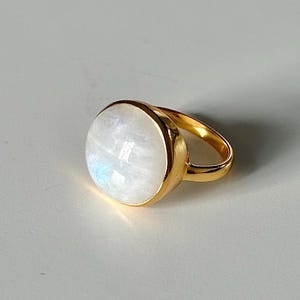Op de afbeelding: Een gouden ring met een grote ovale maansteen. De ring is gezet in een eenvoudige band en de steen is melkachtig wit.