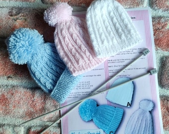 May include: Three knitted baby hats with pom-poms in pink, blue, and white. The hats are laid out on a white sheet of paper with knitting needles and instructions for making the hats. The paper has the text "HolmeCraft" and a bee logo.