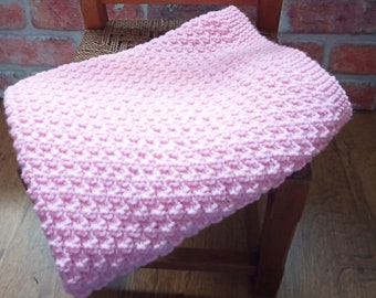 May include: A pink knitted baby blanket with a textured pattern. The blanket is folded and draped over a wooden chair.