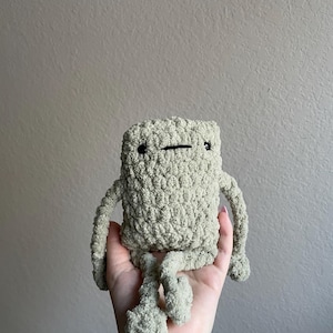 Puede incluir: Un juguete de peluche tejido a crochet con forma de criatura con un cuerpo cuadrado y extremidades largas. El juguete es de color verde claro y tiene ojos negros.