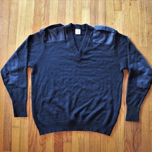 Może przedstawiać: Granatowy sweter z dekoltem w szpic i łatkami na łokciach. Sweter wykonany jest z miękkiej, dzianinowej tkaniny.