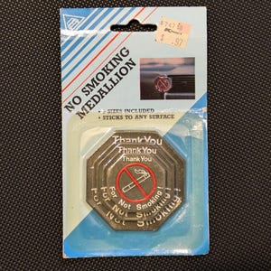 Könnte beinhalten: Ein silberfarbener Metall-Nichtraucher-Medaillon mit dem dreimal wiederholten Text "Danke schön". Das Medaillon zeigt außerdem ein rotes Nichtraucher-Symbol mit einer Zigarette und einer roten Flamme. Der Text "Für Nichtrauchen" ist um den Rand des Medaillons herum geschrieben.