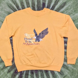 May include: A yellow sweatshirt with a graphic of an eagle flying with a banner that reads "Proud To Be An American".