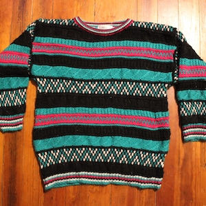 Op de afbeelding: Een gestreepte trui met zwarte, turquoise, roze en witte horizontale strepen. De trui heeft een ronde hals en lange mouwen.
