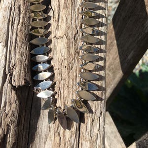 May include: A silver chain necklace with a repeating pattern of leaf-shaped links. The necklace is laying on a piece of driftwood.