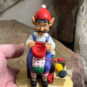 Puede incluir: Una figurita de un elfo con un gorro rojo y un mono azul, sentado y tejiendo una media roja. El elfo tiene barba blanca, pelo negro y gafas. El elfo está sentado sobre una base de madera con una cesta de lana y un carrete de lana roja.
