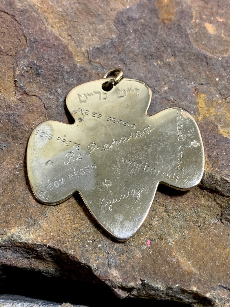 Op de afbeelding: Gouden metalen Girl Scout-speld in de vorm van een klavertjevier. De speld heeft de woorden "Be Prepared" in het Engels, "Soy Prete" in het Spaans, "Sois Prete" in het Frans, "We es Bereid" in het Duits, "EGY Resen" in het Hongaars, "Vant beredt" in het Zweeds en "Be Prepared" in het Hebreeuws. De speld heeft ook een klein klavertjevierontwerp op de achterkant.
