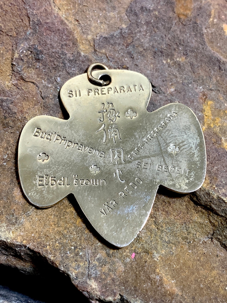Op de afbeelding: Een zilveren metalen Girl Scout-speld in de vorm van een klavertjevier. De speld heeft de woorden "Si Preparata", "Bud' Pripravena", "Sei Bereit", "Var Redo", "Et Bdl'stown" en "Be Prepared" erop.