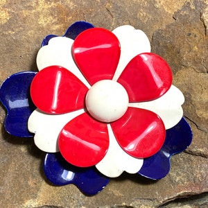 May include: A red, white and blue enamel flower brooch. The flower has six petals and a white centre. The brooch is set against a brown, textured background.