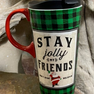 Może przedstawiać: Zielony i czarny kubek podróżny w kratkę z czerwonym uchwytem i czarną pokrywką. Kubek ma białą etykietę z tekstem "Stay jolly my friends" i kreskówkowym obrazkiem Świętego Mikołaja. Tekst "Don't stop believin'" znajduje się również na etykiecie.