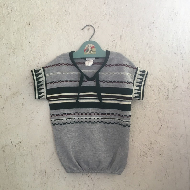 K&ouml;nnte beinhalten: Ein kurz&auml;rmeliges Shirt in Grau und Schwarz mit einem geometrischen Muster an den &Auml;rmeln. Das Shirt hat einen V-Ausschnitt und einen Kordelzug.