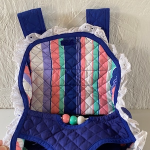 May include: A blue quilted baby carrier with white lace trim and a striped fabric lining. The carrier has two straps and a small pocket on the front. There are three colorful beads hanging from the front of the carrier.