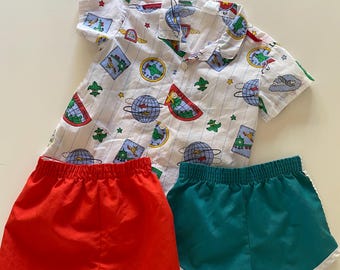 May include: A white short-sleeved shirt with a colorful travel-themed pattern, paired with a pair of red shorts and a pair of teal shorts with white trim. The shirt features images of globes, airplanes, and other travel-related graphics.