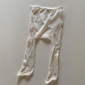 May include: Off-white, open-weave tights. The tights are made of a delicate, textured material. The waistband is a lighter shade of off-white. The legs are long and narrow, with a slight flare at the ankles. The tights are designed for wear.