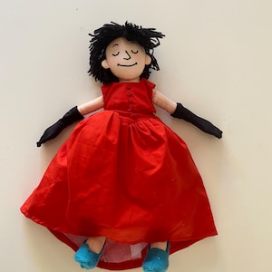 May include: A handmade doll with a smiling face, black hair, and black gloves. The doll wears a red dress and blue shoes. The doll's arms are outstretched, and it appears to be floating against a white background.