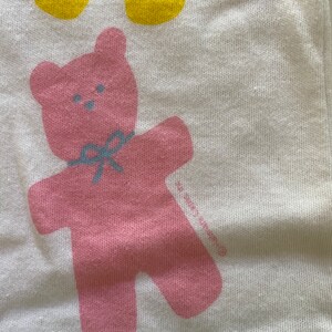 May include: A pink teddy bear with a blue bow is printed on a white background. The bear is standing with its arms outstretched. The background is a light green colour.