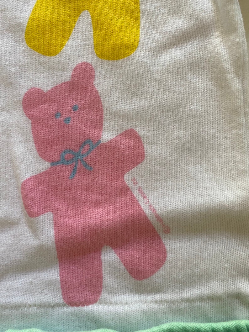 May include: A pink teddy bear with a blue bow is printed on a white background. The bear is standing with its arms outstretched. The background is a light green colour.