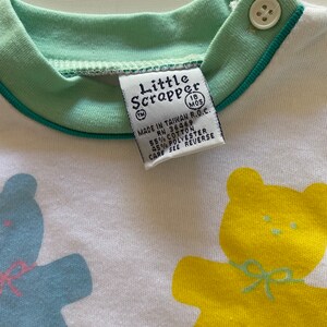 May include: A white baby onesie with a green collar and blue trim. The onesie features two yellow teddy bears with green bows. The text "Little Scrapper" is printed on the onesie with the trademark symbol and the text "18 MOS" in a circle. The text "MADE IN TAIWAN R.O.C. RN 36469 55% COTTON 45% POLYESTER CARE SEE REVERSE" is printed on the onesie.