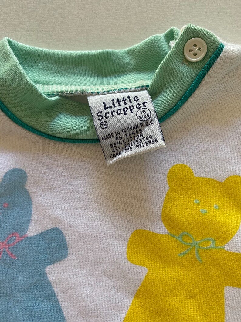 May include: A white baby onesie with a green collar and blue trim. The onesie features two yellow teddy bears with green bows. The text "Little Scrapper" is printed on the onesie with the trademark symbol and the text "18 MOS" in a circle. The text "MADE IN TAIWAN R.O.C. RN 36469 55% COTTON 45% POLYESTER CARE SEE REVERSE" is printed on the onesie.