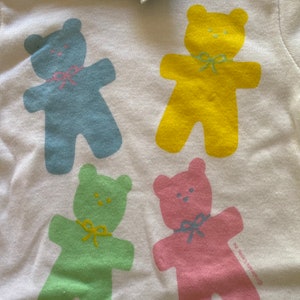 May include: White t-shirt with four colourful teddy bear illustrations. The bears are blue, yellow, green, and pink. The shirt has a green trim around the neck. The text "Little Scrappers" is printed on the shirt.