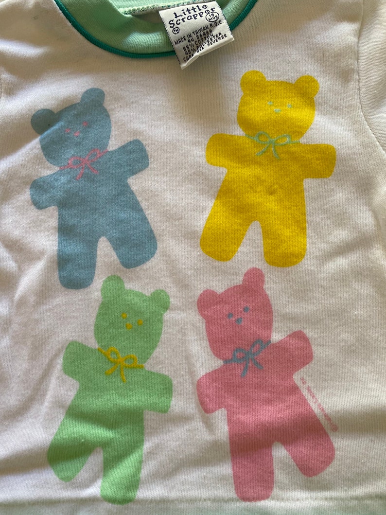 May include: White t-shirt with four colourful teddy bear illustrations. The bears are blue, yellow, green, and pink. The shirt has a green trim around the neck. The text "Little Scrappers" is printed on the shirt.