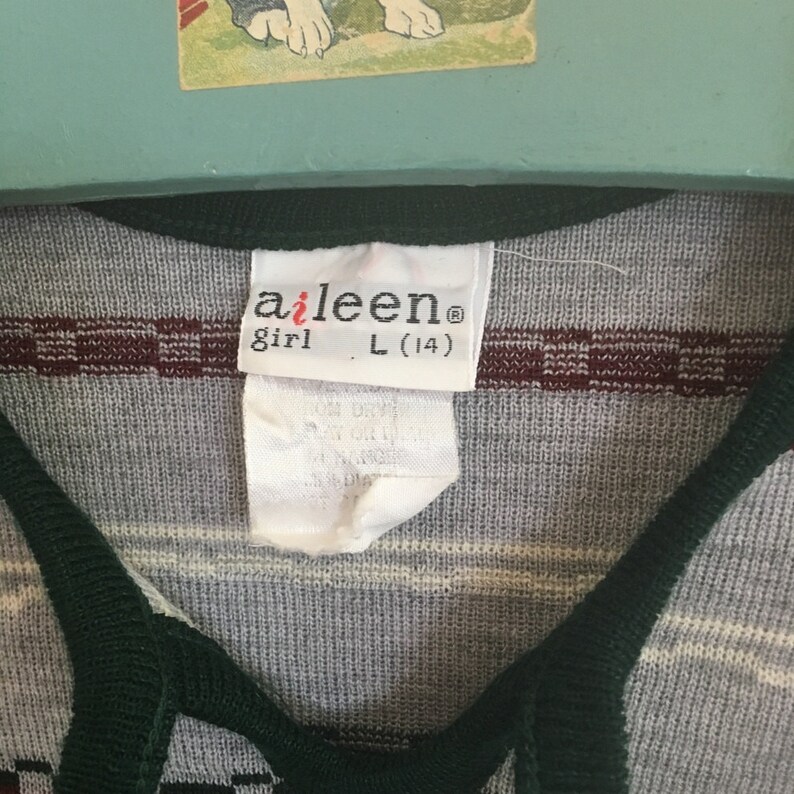 K&ouml;nnte beinhalten: Ein wei&szlig;es Etikett mit dem Text "aileen girl L (14)" auf einem gestreiften Shirt mit gr&uuml;nen und burgunderfarbenen Streifen.