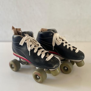 May include: A pair of black high-top roller skates featuring white laces, a thin red stripe along the side of the sole, and four light-colored wheels on each skate. The wheels are marked with the word Syndicate in red lettering on the side.