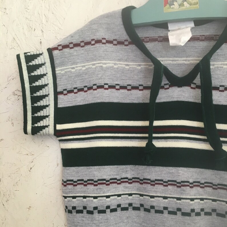 K&ouml;nnte beinhalten: Ein kurz&auml;rmeliger, gestreifter Pullover mit einem geometrischen Muster an den &Auml;rmeln. Der Pullover ist in den Farben Gr&uuml;n, Wei&szlig; und Grau gehalten und hat einen Knoten am Hals.