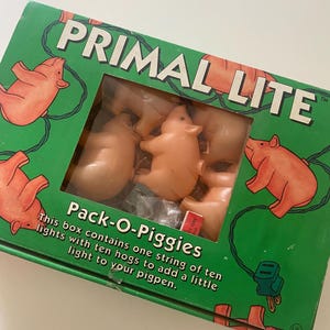 May include: A green box labeled "PRIMAL LITE" with a window showing several small, pink pig figurines. The box also says "Pack-O-Piggies" and includes text about the product. The box has illustrations of pigs.