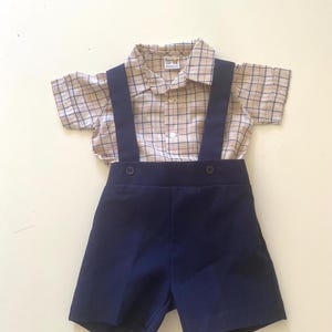 May include: A vintage-style baby outfit featuring a plaid short-sleeved shirt and navy blue shorts with suspenders. The shirt has a button-down collar and the shorts have two buttons on the front. The outfit is designed for a baby or toddler.